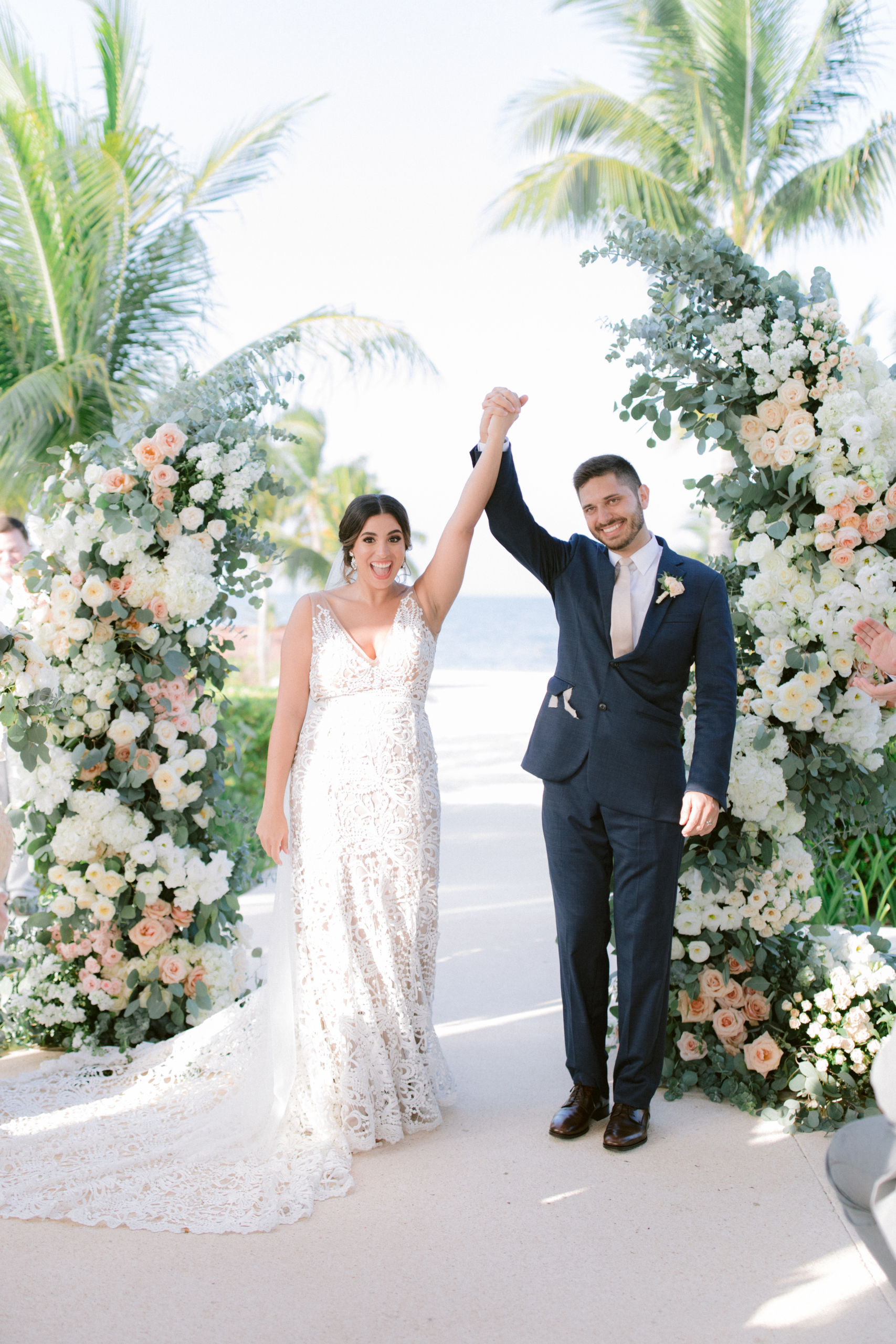 Newlyweds standing at a floral arch with palmtrees in the background, hold hands and raise them in the air to celebrate that they are just married.
