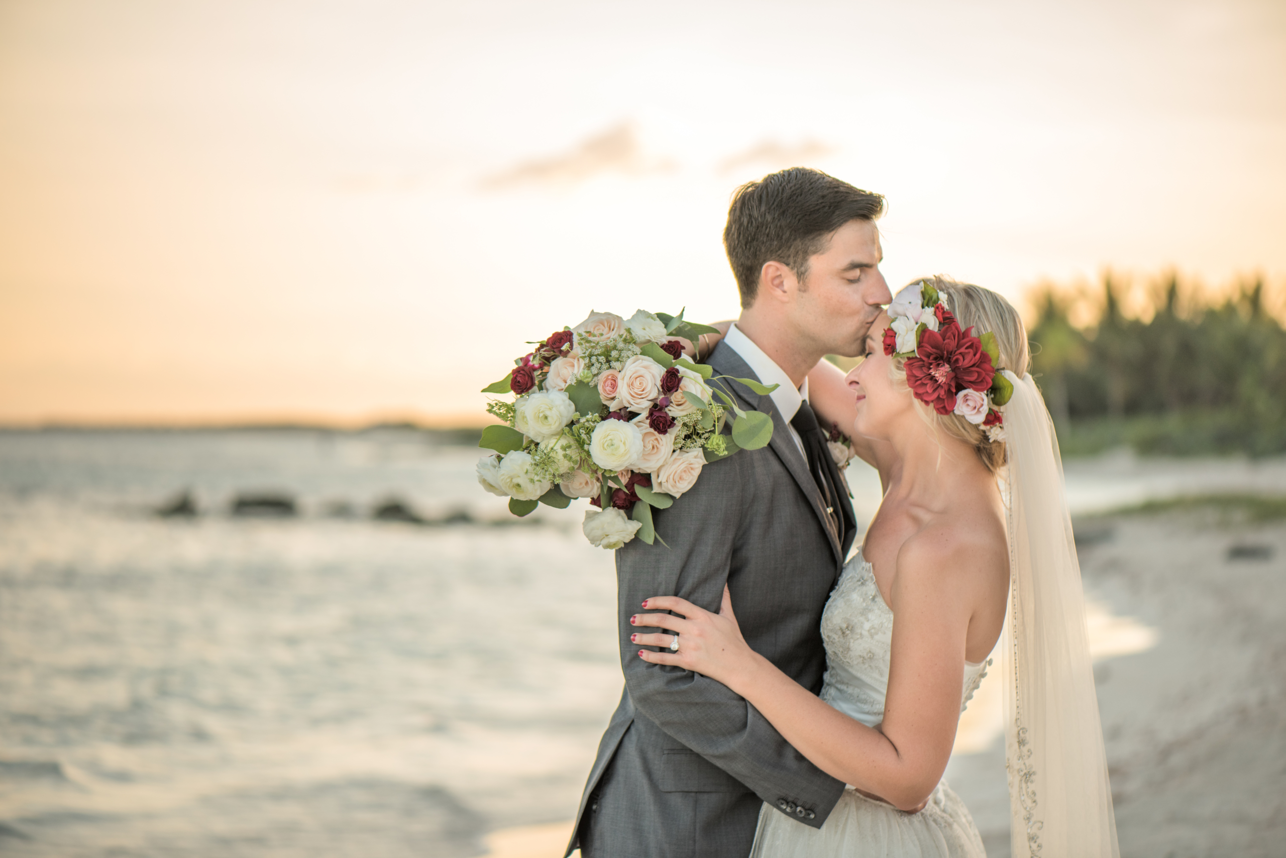 Groom kisses his new bride on the forehead as they embrace on the beach in Tulum with the sun setting in the background. The bride holds a beautiful bouquet