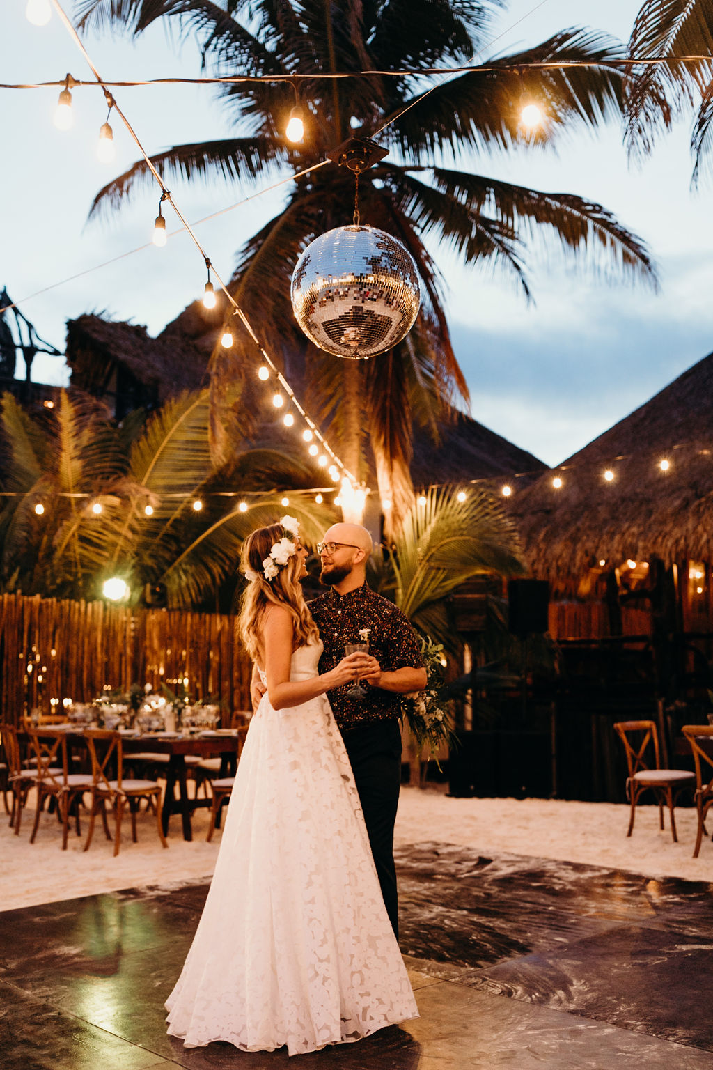 Bride and Groom share their first dance on a wood dance floor under a glimmering disco ball and string lights, Plam trees in the background at Akiin Beach Wedding